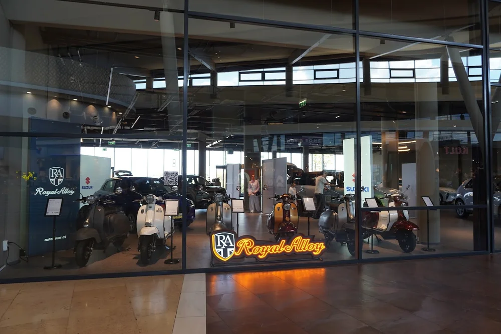 Royal Alloy Motorroller in verschiedenen Farben im Schaufenster des Auto Auer Popup Stores im G3, im Hintergrund Suzuki Fahrzeuge